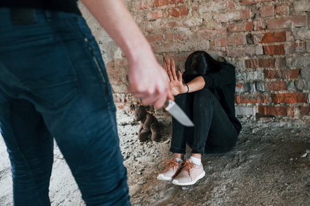 Violent man standing with knife in hand and threatens girl that sits on the floor in abandoned buildingの写真素材