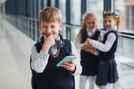 Little boywith phone and headphones standing if front of school kids in uniform that together in corridor. Conception of educationの写真素材