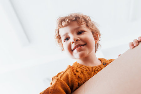 View from below of little boy with curly hair that have fun with paper box indoorsの写真素材