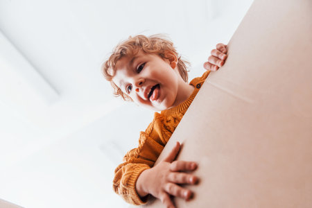 View from below of little boy with curly hair that have fun with paper box indoorsの写真素材