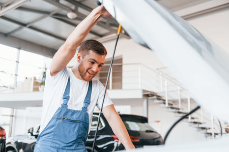 Young man in white shirt and blue uniform repairs automobileの写真素材