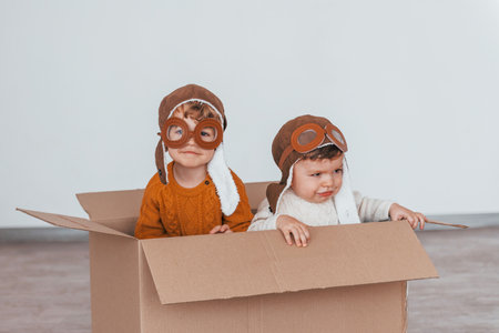 Two little boys in retro pilot costumes have fun and sitting in paper box indoors at daytimeの写真素材