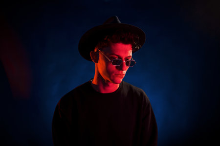 In hat and glasses. Neon lighting. Young european man is in the dark studioの写真素材