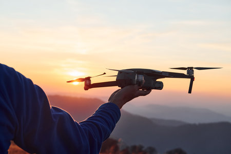 Man standing in the autumn field and holds drone in hand. Beautiful sunrise. Majestic landscape of mountains far awayの写真素材