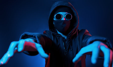 In mask, eyewear and hood. Neon lighting. Young european man is in the dark studioの写真素材