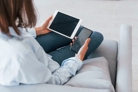 Holds smartphone and tablet. Young woman is indoors in room of modern house at daytimeの写真素材