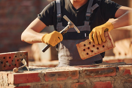 Holding brick and using hammer. Construction worker in uniform and safety equipment have job on...