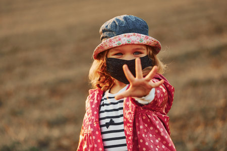 Showing hand. Portrait of cute little girl in black protective mask that standing outdoorsの写真素材