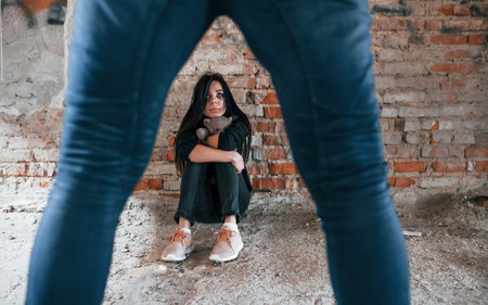 Violent man standing and threatens girl that sits on the floor with teddy bear in abandoned buildingの写真素材