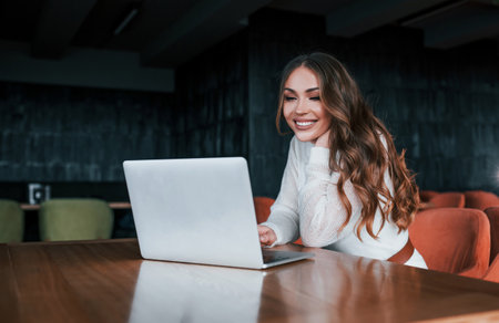 Successful young woman in white dress sits by table with laptopの写真素材