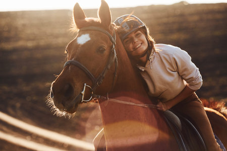 Young woman in protective hat with her horse in agriculture field at sunny daytime