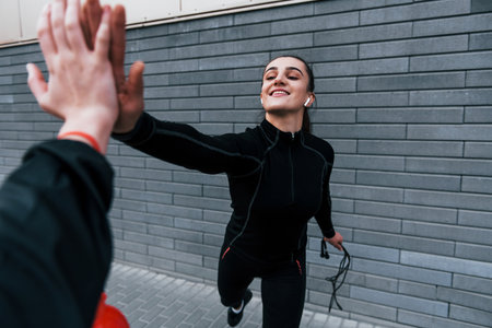 Young sportive girl in black sportswear standing with jump rope in hands outdoors near gray wall and giving high fiveの写真素材