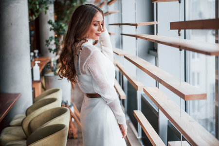 Gorgeous woman in white dress standing indoors near windowsの写真素材
