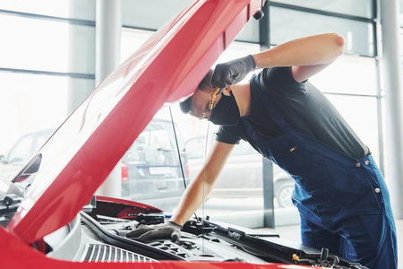 Man in uniform and black protective mask works with broken automobile. Conception of car serviceの写真素材