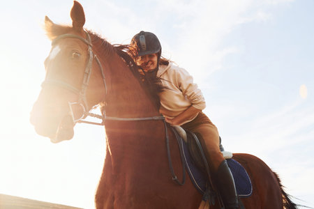 Young woman in protective hat with her horse in agriculture field at sunny daytime