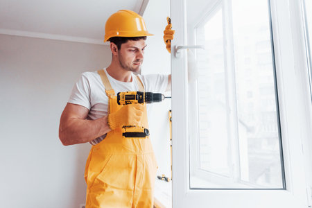 Handyman in yellow uniform installs new window by using automatic screwdriver. House renovation conceptionの写真素材