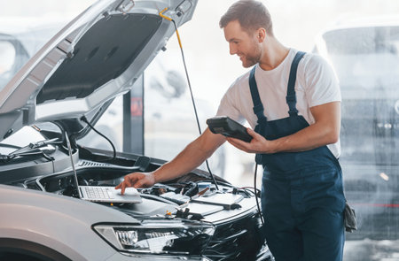 Young man in white shirt and blue uniform repairs automobileの写真素材