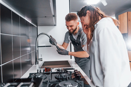 Young professional plumber in grey uniform shows results of work for housewife on the kitchenの写真素材