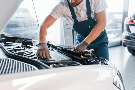 Quality service. Young man in white shirt and blue uniform repairs automobileの写真素材