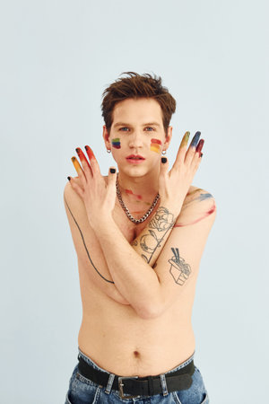 Young gay man is standing in the studio and posing for a camera. Shirtless guy with rainbow make up