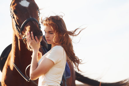 Beautiful sunshine. Young woman standing with her horse in agriculture field at daytimeの写真素材