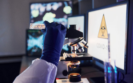 Scientist in uniform and gloves holds lens with COVID-19 title on it. Indoors in laboratory