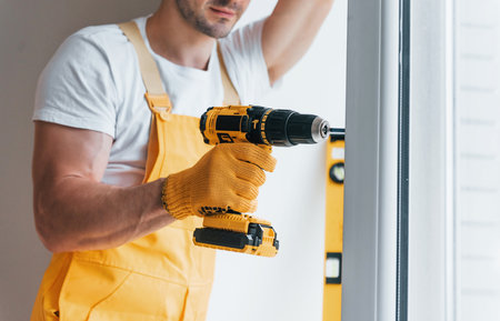 Handyman in yellow uniform installs new window by using automatic screwdriver. House renovation conceptionの写真素材