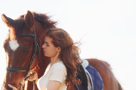 Beautiful sunshine. Young woman standing with her horse in agriculture field at daytimeの写真素材