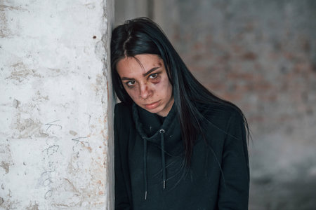 Portrait of beaten young woman with bruise under eye that standing and leaning on the wall indoors...