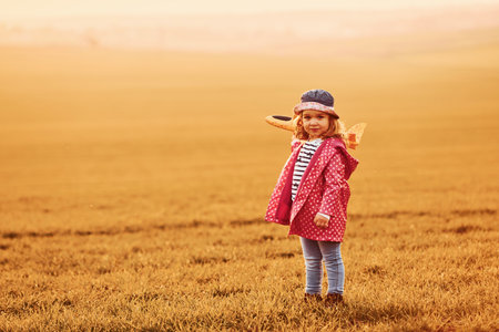 Illuminated by orange colored sunlight. Cute little girl have fun with toy plane on the beautiful field at daytimeの写真素材