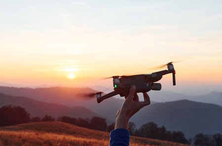 Man standing in the autumn field and holds drone in hand. Beautiful sunrise. Majestic landscape of mountains far awayの写真素材