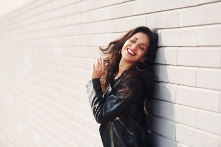 Potrait of cheerful brunette with curly hair and in black clothes leaning on the wallの写真素材