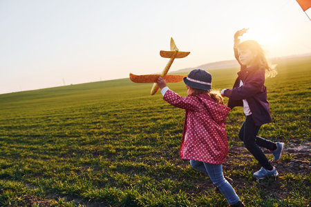 Two little girls friends have fun together with kite and toy plane on the field at sunny daytimeの写真素材