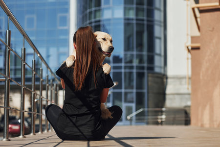 Sitting together. Young positive woman have fun with her dog when have a walk outdoors near business buildingの写真素材