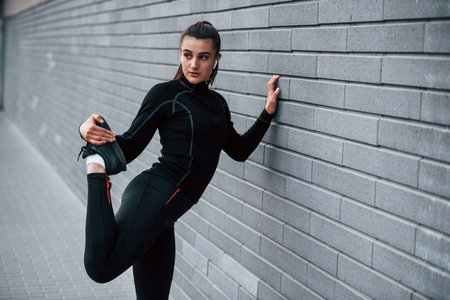 Young sportive girl in black sportswear outdoors doing stretching near gray wall