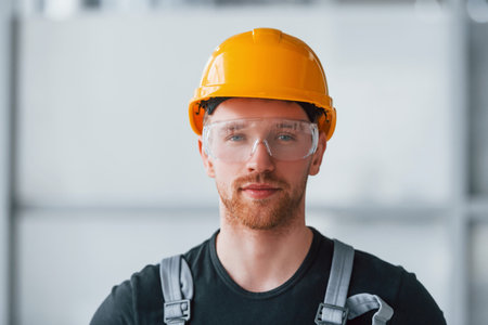 Portrait of man in grey uniform, protective glasses and orange hard hat that standing indoors in modern big office at daytimeの写真素材