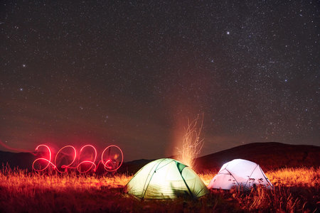 2020 neon numbers. Two iluminated tents with campfire under stars at mountains at nightの写真素材