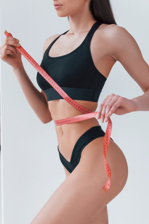 Slender woman in black fitness underwear standing indoors in room at daytime with measuring tapeの写真素材