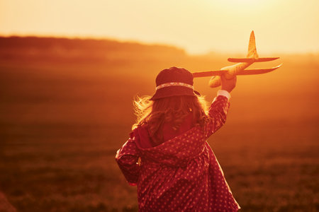 Illuminated by orange colored sunlight. Cute little girl have fun with toy plane on the beautiful field at daytimeの写真素材