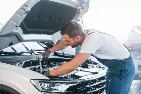Quality service. Young man in white shirt and blue uniform repairs automobileの写真素材