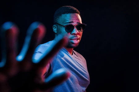 With sunglasses. Futuristic neon lighting. Young african american man in the studioの写真素材
