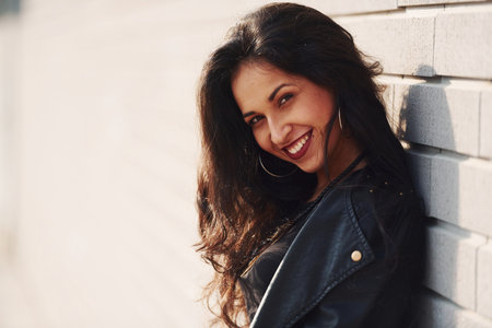 Potrait of cheerful brunette with curly hair and in black clothes leaning on the wallの写真素材