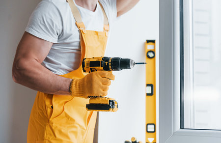 Handyman in yellow uniform installs new window by using automatic screwdriver. House renovation conceptionの写真素材