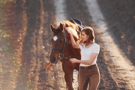 Young woman in protective hat walking with her horse in agriculture field at sunny daytime