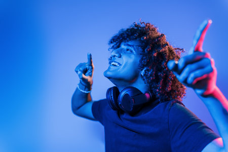 With headphones. Young beautiful man with curly hair is indoors in the studio with neon lightingの写真素材