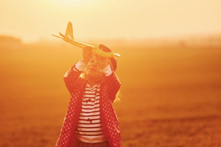 Illuminated by orange colored sunlight. Cute little girl have fun with toy plane on the beautiful field at daytimeの写真素材