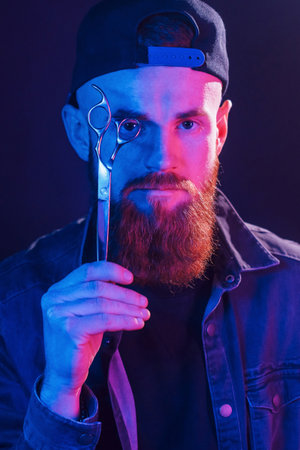 With scissors. Young bearded barber in cap standing in the studio with neon lightingの写真素材