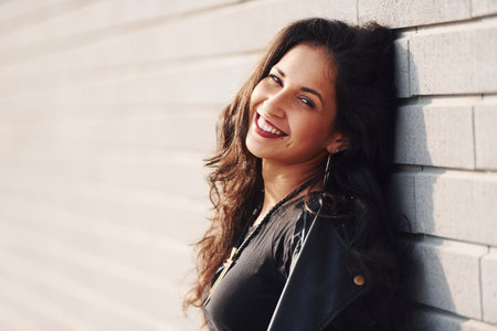 Potrait of cheerful brunette with curly hair and in black clothes leaning on the wallの写真素材