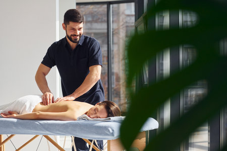 Room illuminated by sun beams. Young woman is lying down when man doing massage of her body at spaの写真素材