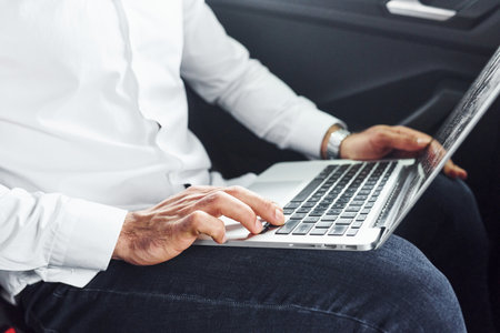 Man in white shirt is sitting inside a car with laptop that is on his legsの写真素材